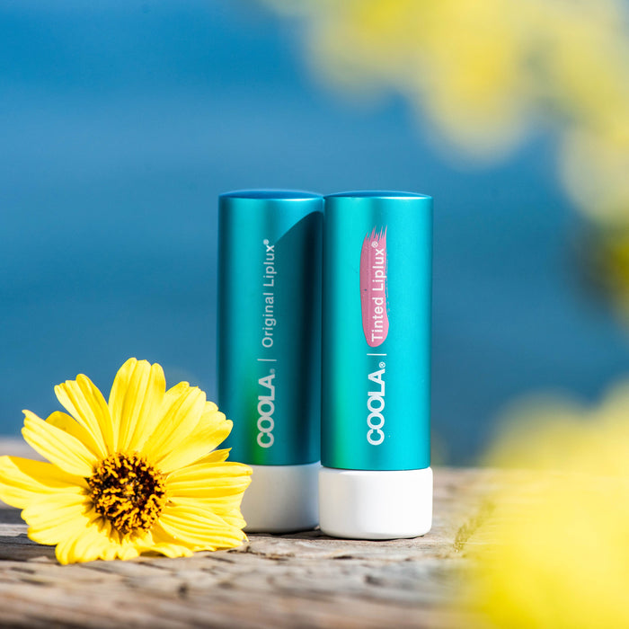 Two teal lip balm tubes labeled COOLA Original Liplux and Tinted Liplux placed on a wooden surface near yellow flowers, showcasing Classic Liplux® Lip Balm Tinted SPF 30 | COOLA for skincare and sun protection.