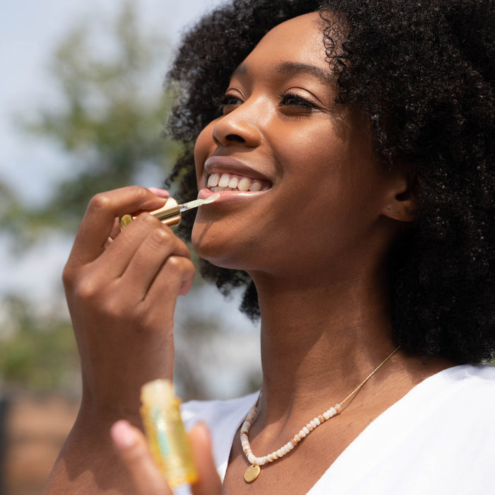 Woman outdoors smiling while applying Classic Liplux® Organic Hydrating Lip Oil Sunscreen SPF 30, showcasing skincare and sun protection in a natural setting.