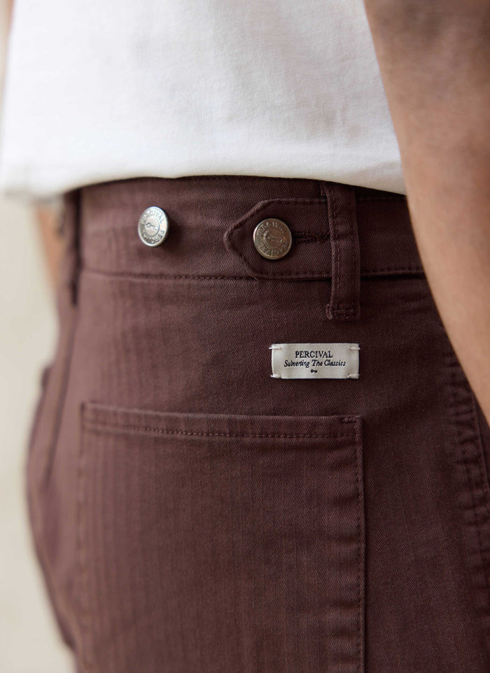Close-up of Percival espresso brown cotton herringbone trousers, featuring branded silver hardware and a woven logo tag.