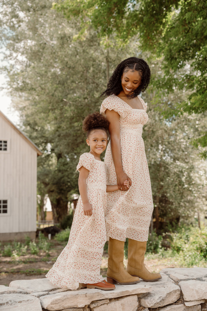 A woman and young girl, both wearing matching floral dresses with smocked bodices, stand hand-in-hand on a stone ledge, smiling. Behind them, green trees and a white barn complete the scene. The girl’s charming cotton dress is the Mini Mirabelle Dress in Cream Ditsy Floral by Ivy City Co.