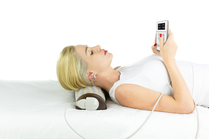 Woman lying on a bed using the Amethyst Bolster Firm - Heated InfraMat Pro® | HealthyLine with a handheld remote for wellness and recovery therapy.