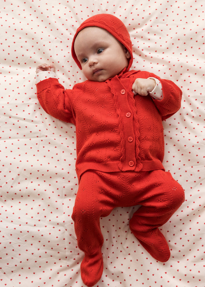 Baby wearing a red cardigan, pants, and matching hat.
