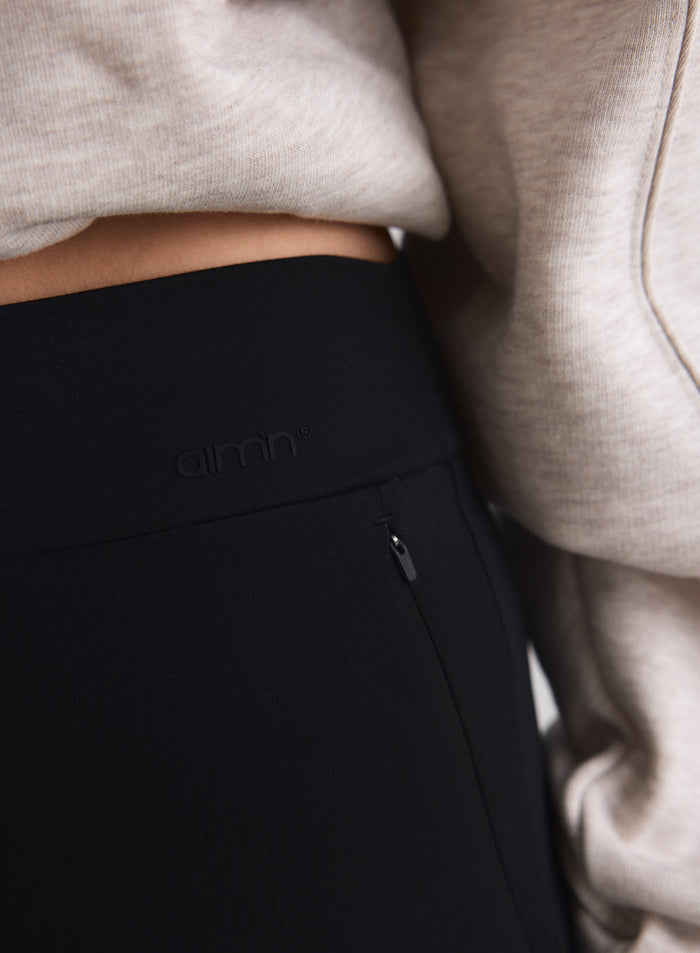 Close-up of black aim'n Sense Pants waistband with a discreet zipped pocket and logo detail, paired with a beige top [M-170cm]