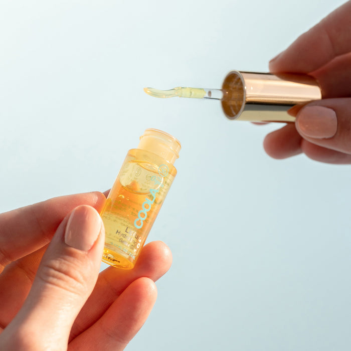 Hand holding a small bottle of Classic Liplux® Organic Hydrating Lip Oil Sunscreen SPF 30 with a dropper applicator lifting clear golden oil against a light background in a skincare setting.
