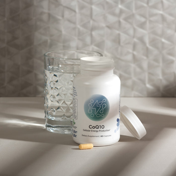 nfiniWell CoQ10 supplement bottle with open lid, capsule, and glass of water on a tabletop.