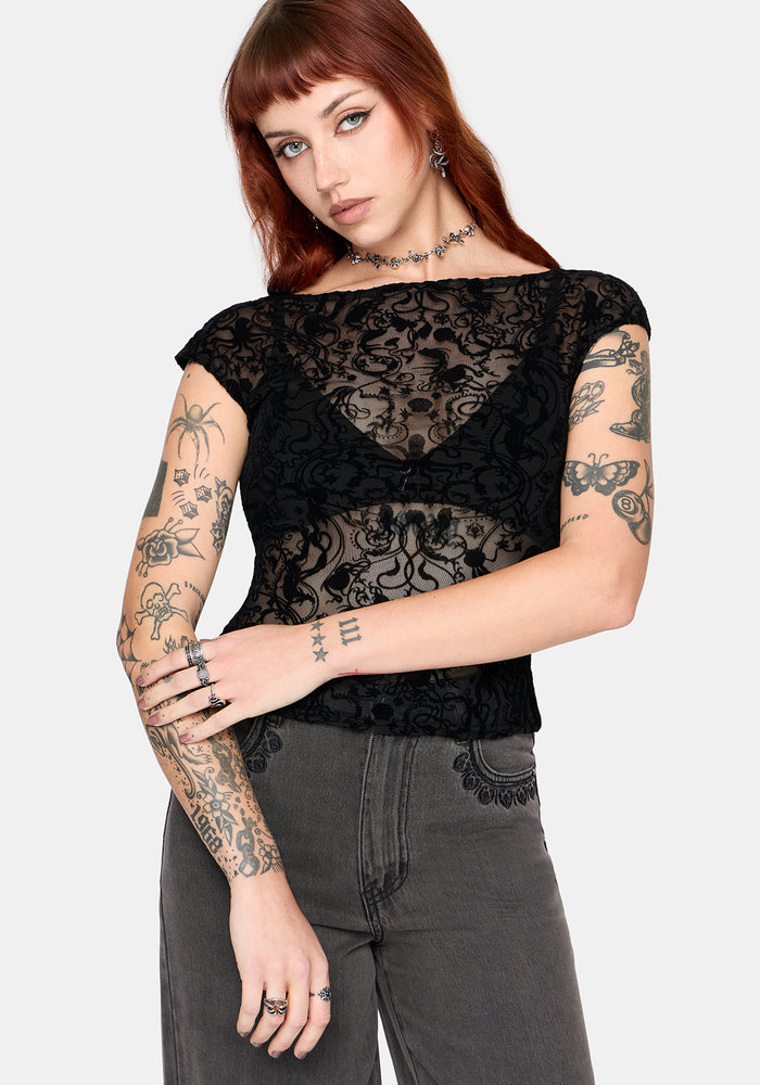 Woman wearing a sheer black lace top and gray pants with tattoos on arms and hands.