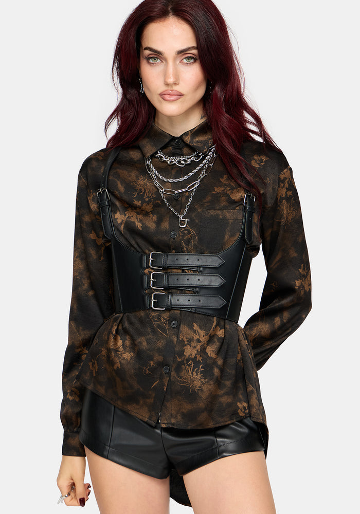 A model with dark red hair wearing a black and brown floral patterned long-sleeve blouse with a black faux-leather corset-style waist belt, layered silver necklaces, and black faux-leather shorts, set against a plain light gray background.