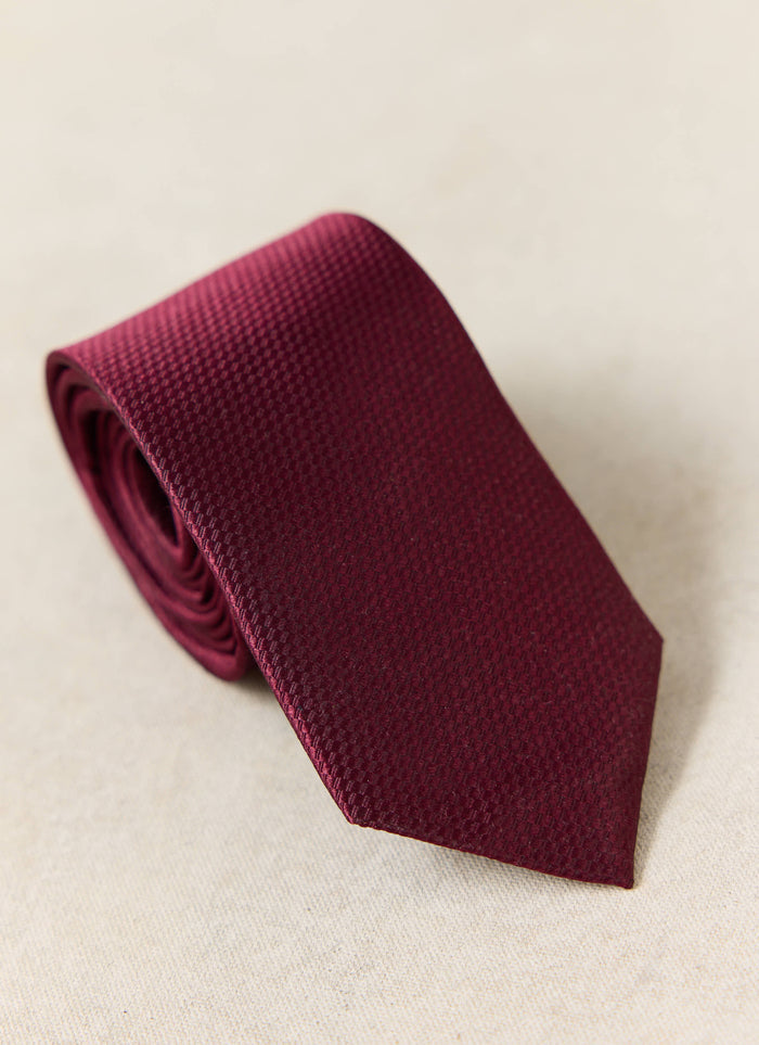 The Percival Grenadine Tie in Red Silk, featuring a textured weave, shown rolled up on a neutral background.