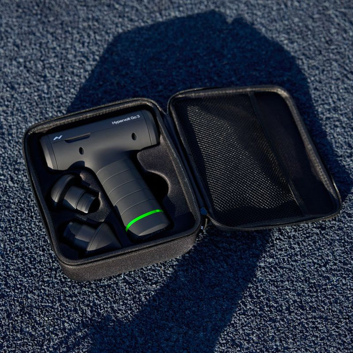 Compact black massage device with three interchangeable heads neatly stored in an open black carrying case on a textured blue surface labeled Hypervolt Go 3 | Hyperice.