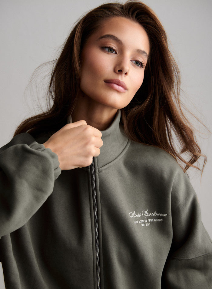 Close-up of a woman in an olive green aim'n Script Zip Jacket with a high collar and logo. [S-174cm]