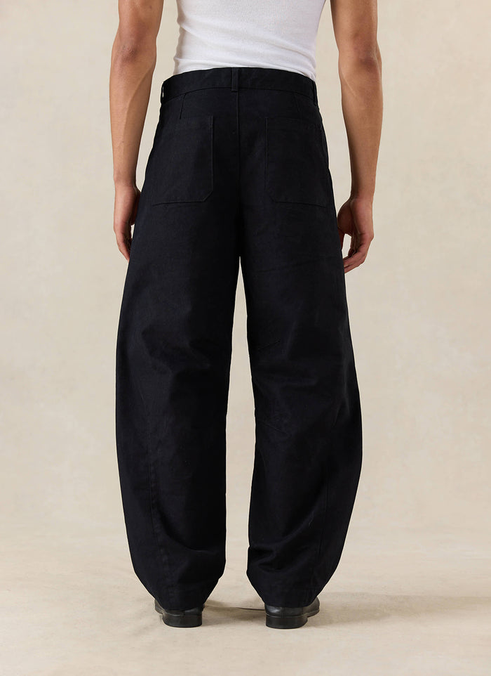 A model wears the Percival Milo Relaxed Barrel Leg Trousers in black, shown from the rear and styled with a white vest.