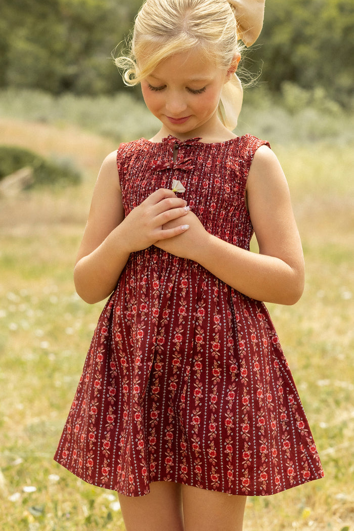 A young blonde girl in the Ivy City Co Mini Ana Dress in Sequoia Felicity Floral stands outside on grass, looking down with her hands together, surrounded by trees and greenery in the blurred background.