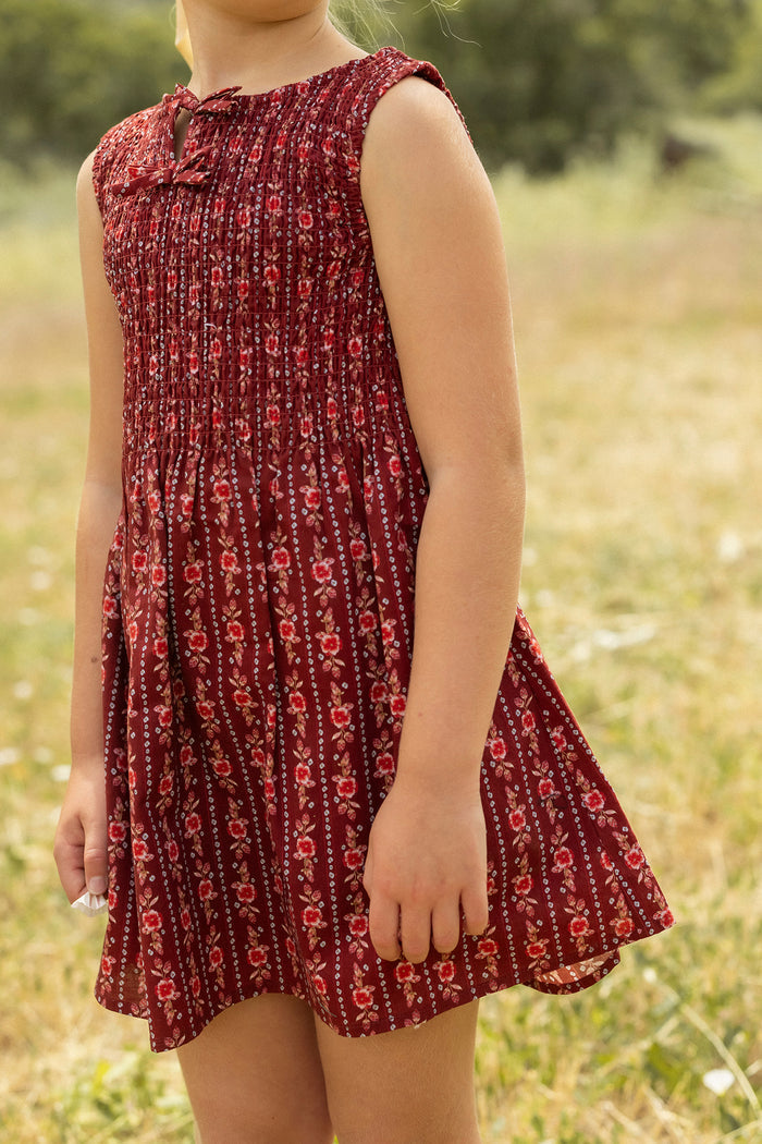 A young girl stands in a grassy field wearing the Ivy City Co Mini Ana Dress in Sequoia Felicity Floral, a red sleeveless style with gentle bodice gathers. Her face is out of frame, and her arms hang relaxed at her sides.