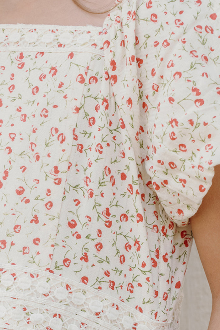A close-up of the Mini Mirabelle Dress in Cream Ditsy Floral by Ivy City Co—a white fabric blouse featuring a red floral pattern with green stems, puffed short sleeves, delicate lace trim, and a glimpse of a person’s shoulder and upper arm—highlighting the charming smocked bodice detailing signature to this design.