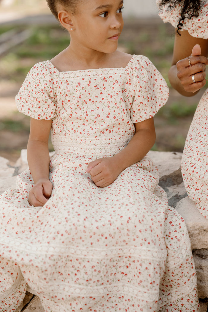 A young girl wears the Ivy City Co Mini Mirabelle Dress in Cream Ditsy Floral, featuring a smocked bodice, as she sits on a stone bench outdoors. Beside her sits an adult with only her arm and hand visible, wearing a matching dress.