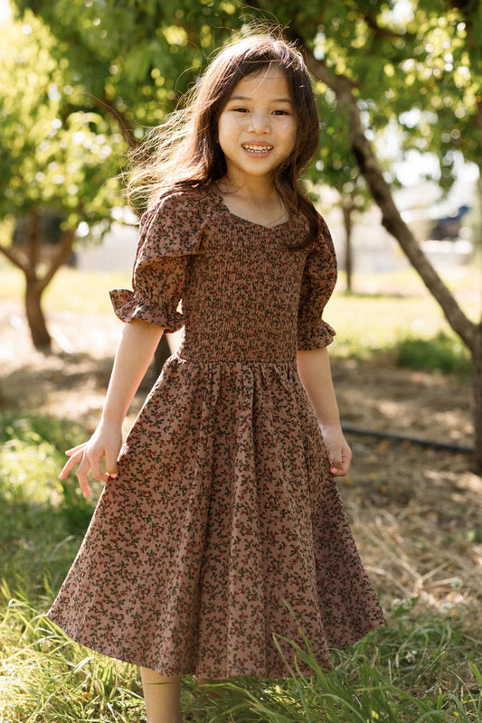 A young girl stands smiling outdoors on a sunny day, surrounded by green trees and grass, wearing the Ivy City Co Mini Teagan Dress in Mauve Woodland Floral with a smocked bodice.