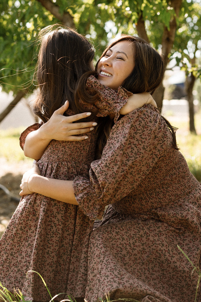 A woman and a young girl, both wearing the Ivy City Co Mini Teagan Dress in Mauve Woodland Floral with charming smocked bodices, hug each other and smile outdoors under leafy trees.