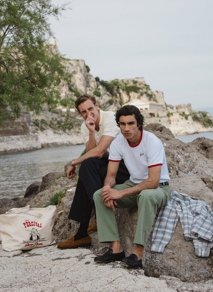 Percival white ringer t-shirt and sage trousers on a model by the sea; check out the full SS26 collection.