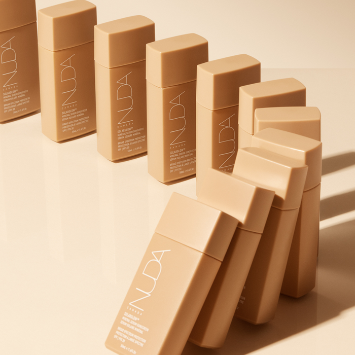 Multiple bottles of Solarglow™ Mineral Serum Sunscreen | NUDA in sleek beige packaging arranged on a reflective surface, highlighting skincare product design and wellness care.