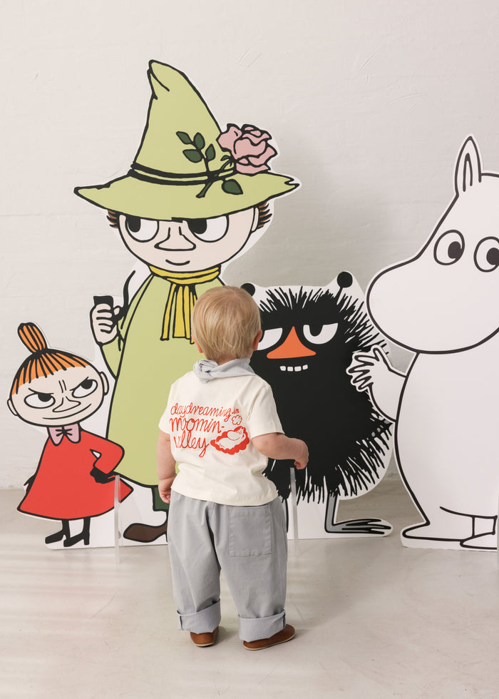 Baby wearing a white T-shirt with Mumin print and light pants together with Moomin figures.