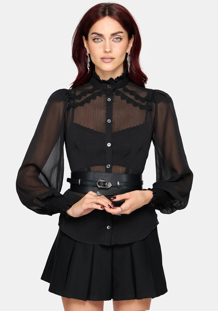 A woman modeling a black long-sleeve blouse with sheer mesh sleeves, a high ruffled collar, and a sheer yoke panel, paired with a matching pleated black skirt and a black belt with silver hardware, against a light gray background.