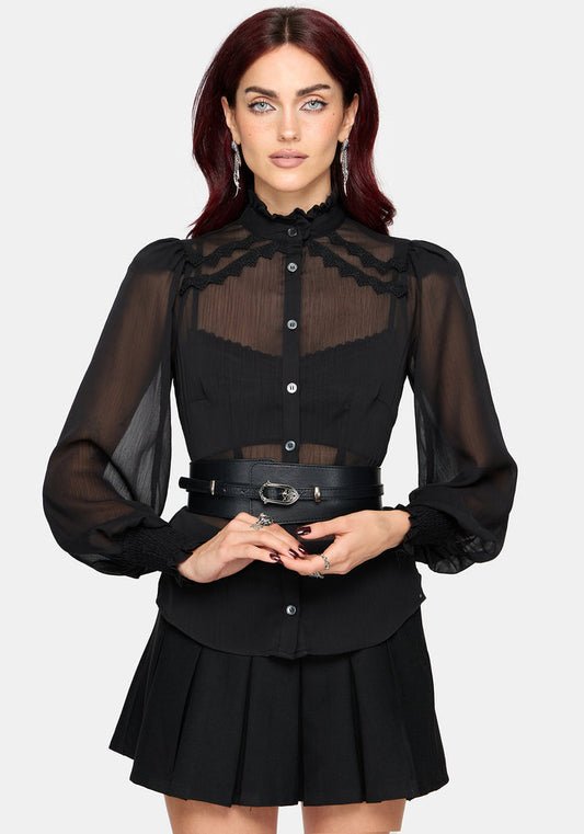 A woman modeling a black long-sleeve blouse with sheer mesh sleeves, a high ruffled collar, and a sheer yoke panel, paired with a matching pleated black skirt and a black belt with silver hardware, against a light gray background.